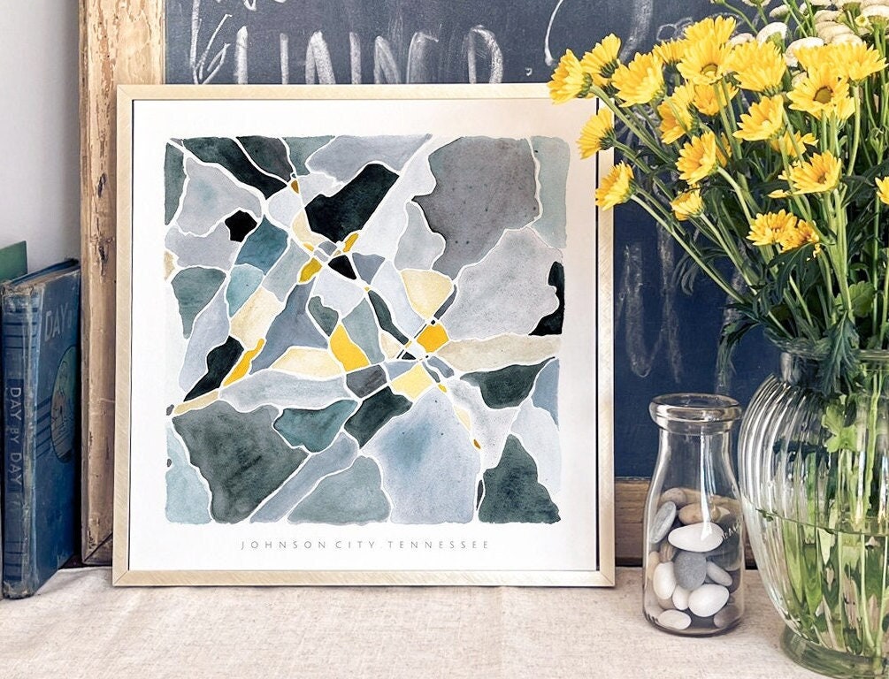 A blue and yellow map of Johnson City, TN in a gold frame beside a vase of yellow flowers, a small vase of smooth stones, and some vintage books.