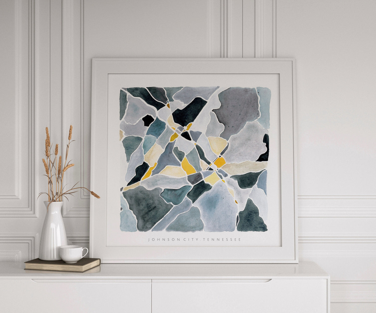 A blue and yellow map of Johnson City, Tennessee in a white frame on a white cabinet beside a book, a vase of wheat and a coffee cup. Behind the picture is an elegant background of white paneling and moulding.