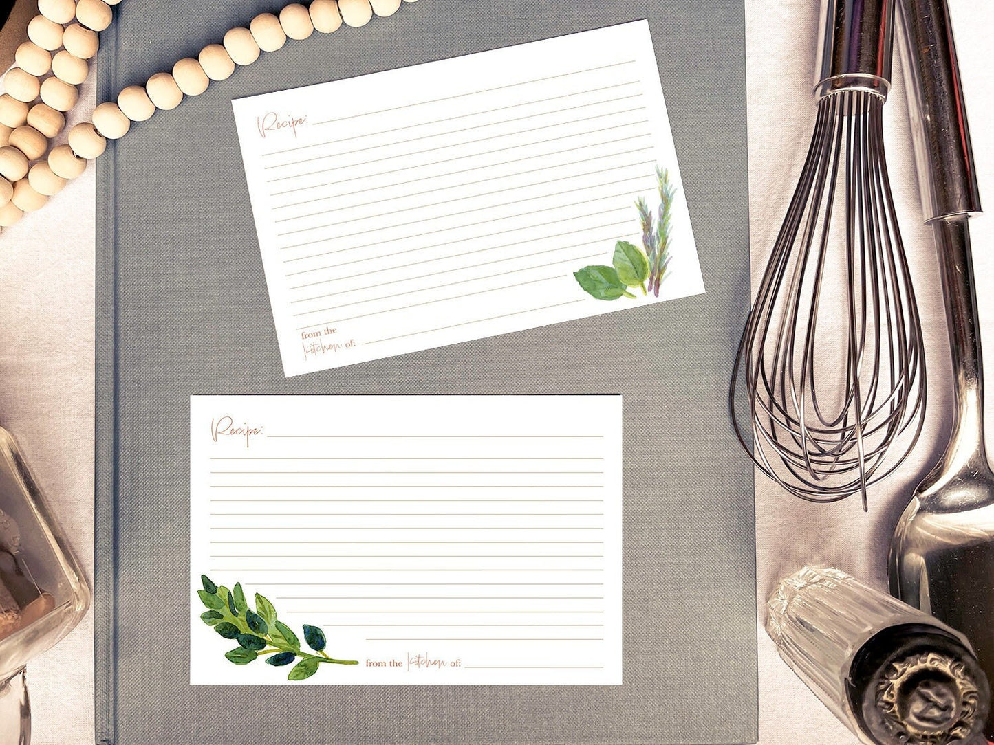 Two hand painted botanical recipe cards atop a grey book surrounded by wooden beads, a silver whisk and spoon, and vintage glass kitchenware.