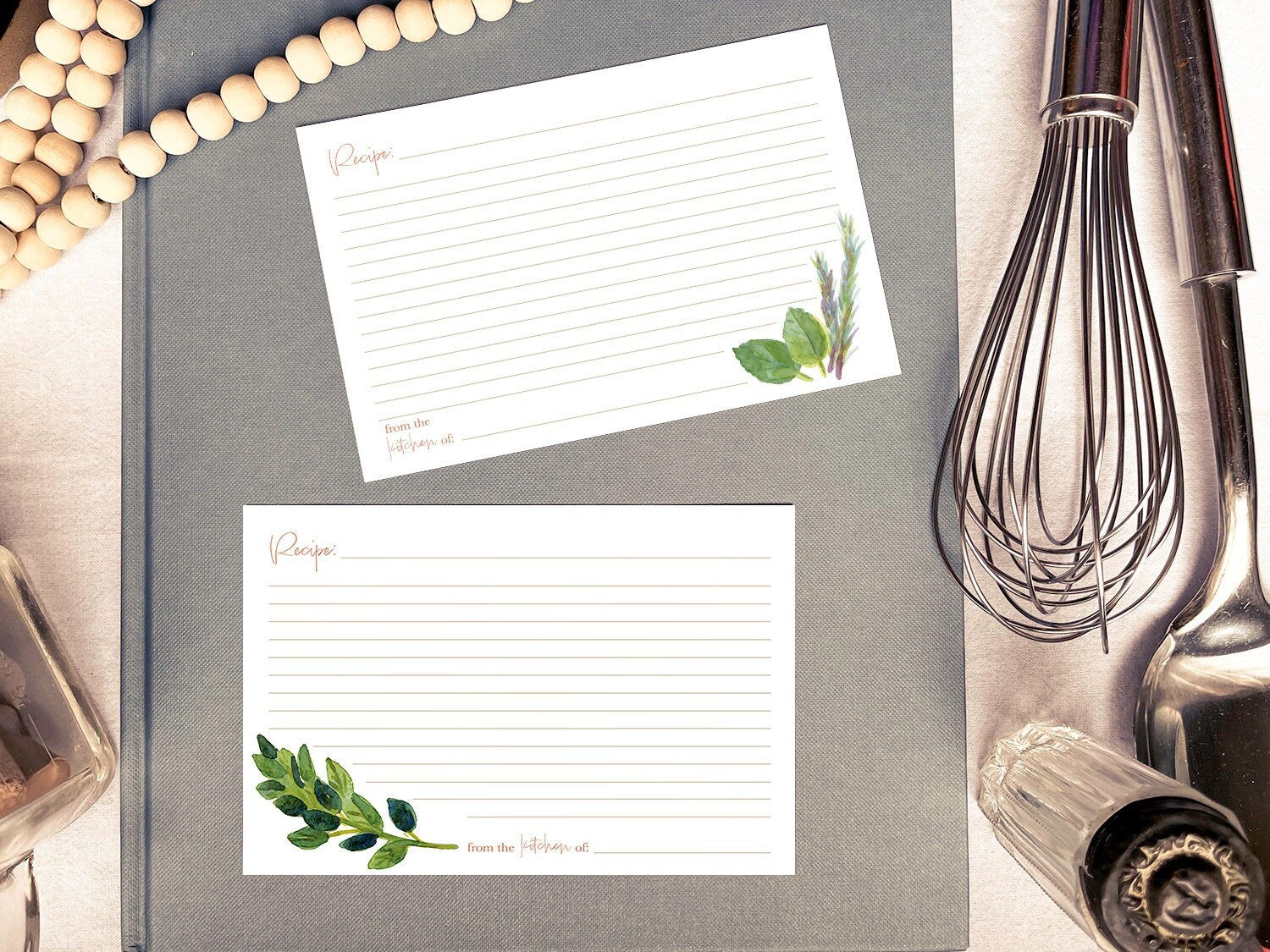 Two hand painted botanical recipe cards atop a grey book surrounded by wooden beads, a silver whisk and spoon, and vintage glass kitchenware.