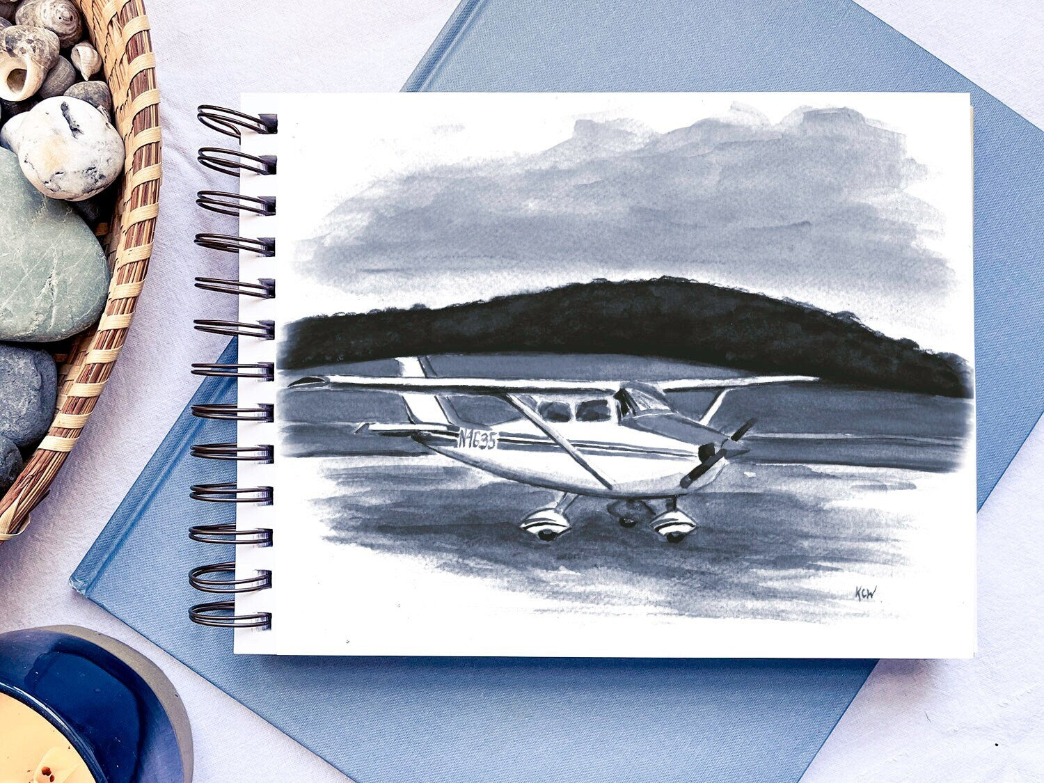 A monochromatic painting of a small airplane in a sketchbook atop a blue book. A basket of rocks and shells and a candle sit nearby.