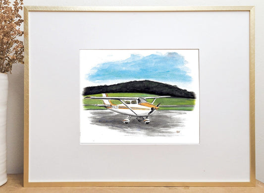 A watercolor illustration of a small yellow airplane in a gold frame near a vase of dried yellow flowers.